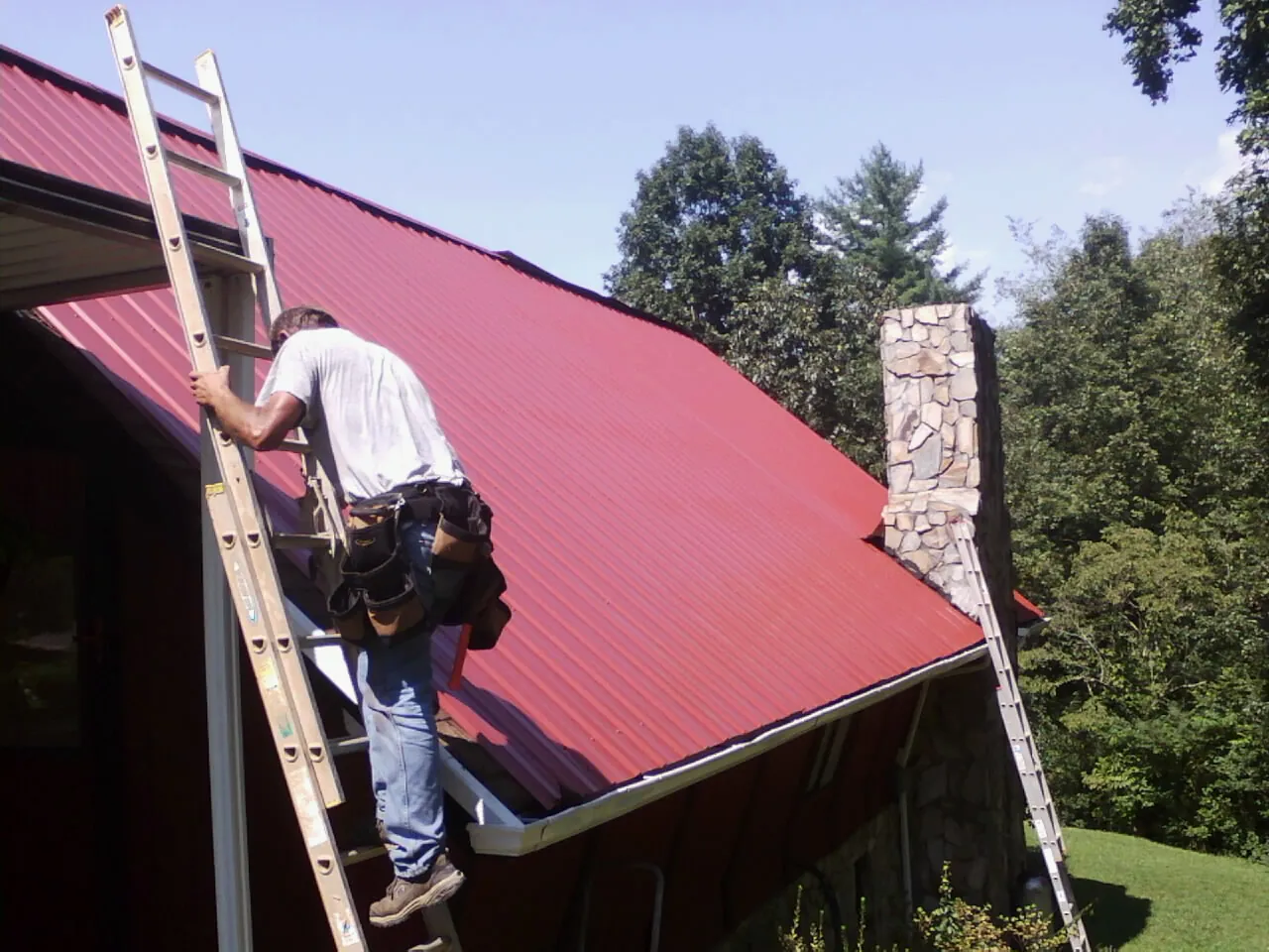 Roofer on a ladder beside a red metal roof and stone chimney — Soffit & Fascia Repair by Roofing Brothers in Concrete